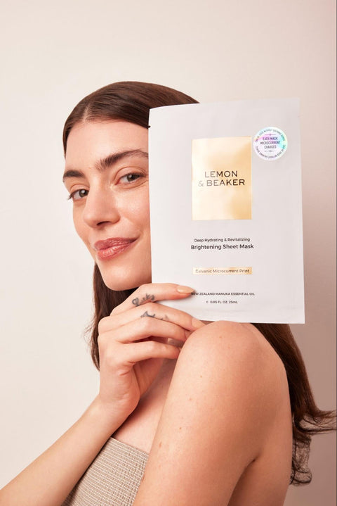 A woman with glowing and healthy skin holds up a Lemon & Beaker Brightening Sheet Mask.