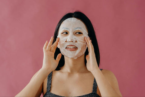 A woman smiles while pressing a sheet mask onto her face.