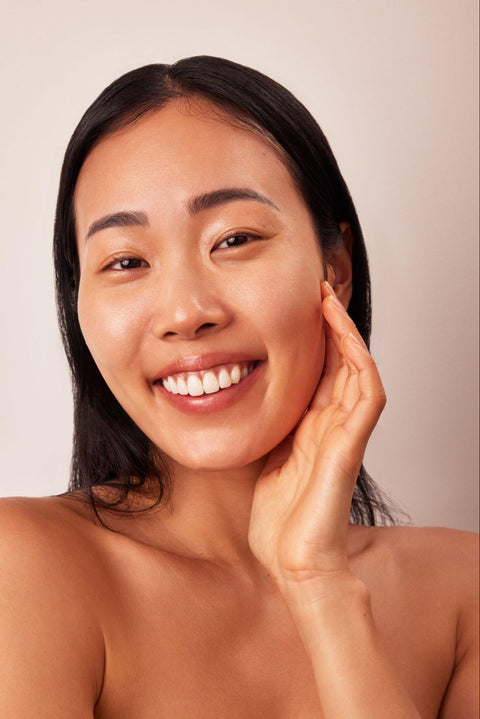 A smiling woman with healthy, glowing skin places a hand on her cheek.