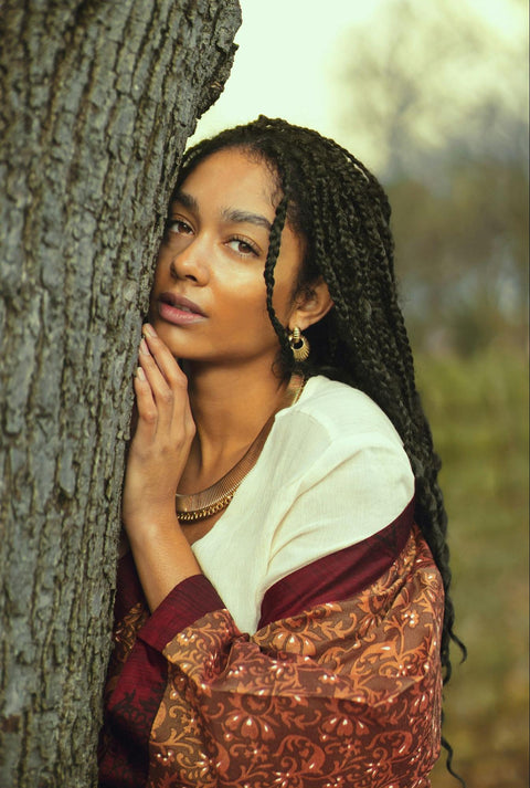 A woman with beautiful skin stands leaning on a tree.