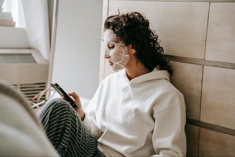 A woman with a sheet mask on her face is looking at her phone as she relaxes.