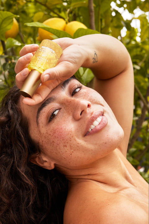 A woman with healthy, glowing skin poses while holding a bottle of serum.