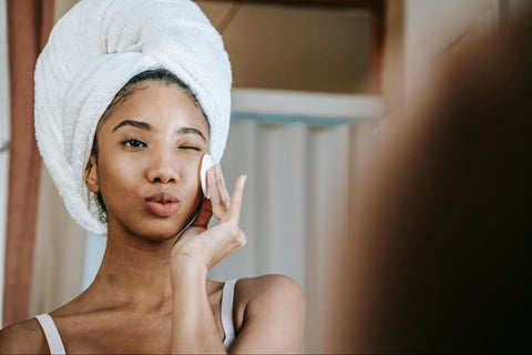 A woman with her hair in a towel uses a cotton pad on her face while winking into the mirror.