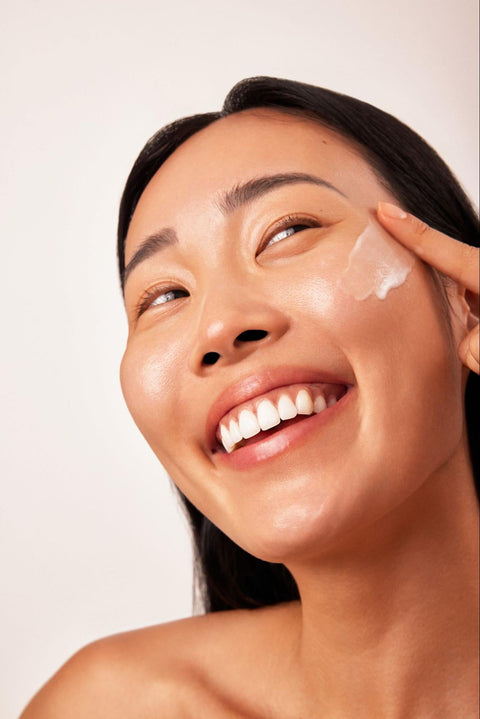 A woman gently applies moisturizer to her cheek while smiling.