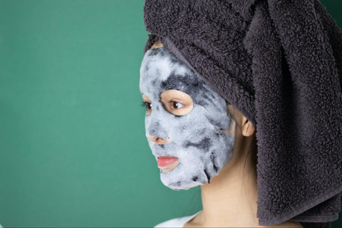 A woman with a towel on her head wears a sheet mask that has light suds on it.