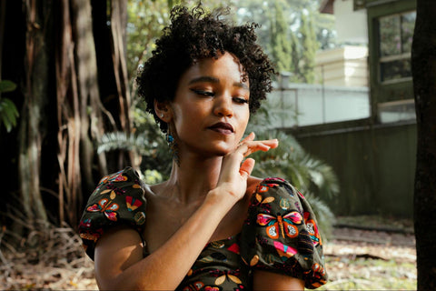 A woman in a colorful top touches her face while sitting among trees with buildings in the background.